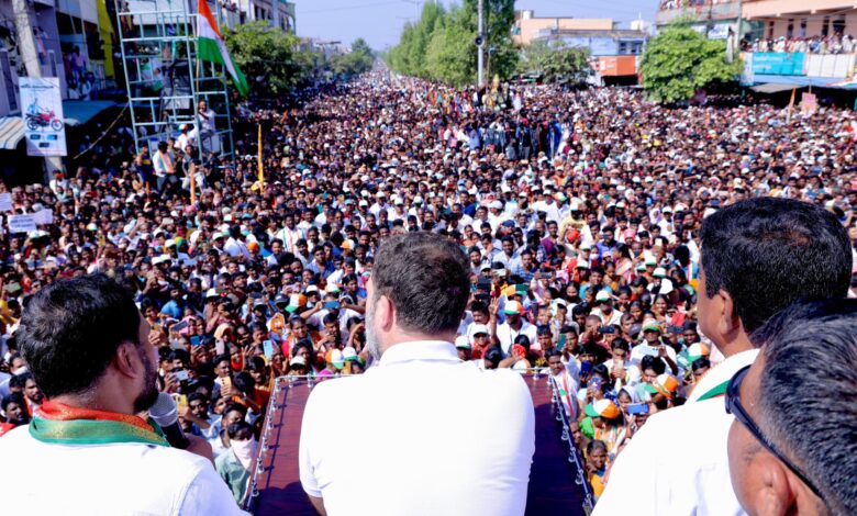 Rahul Gandhi facing a crowd at an election meeting viewed from behind with other Congress leaders on stage.