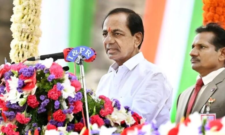 Telangan CM KCR of BRS in white shirt speaking at a government event with flowers in front and flag in back.